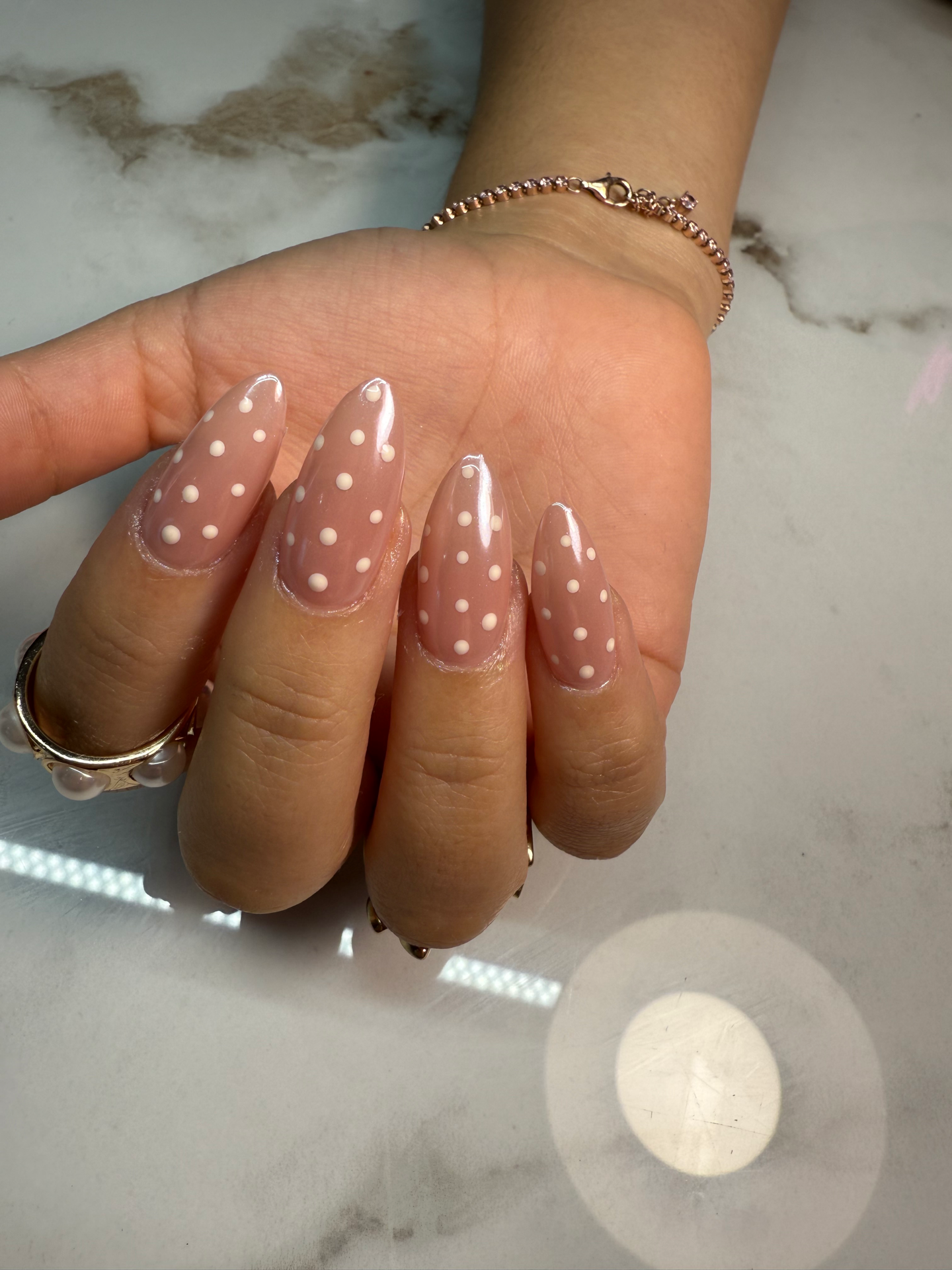 A close-up of a hand with long, almond-shaped nails decorated in a pink base with white polka dots, resting on a light marble surface.