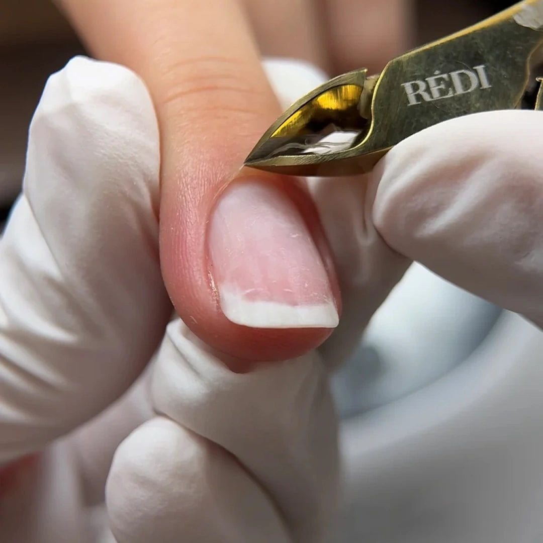 A close-up of a cuticle being clipped with a gold REDI brand manicure tool, held by a hand wearing a white glove.