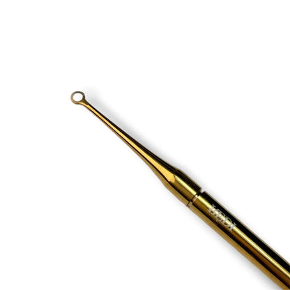 A gold-colored metal scrapping manicure tool with a long, slender handle and a small looped tip, shown diagonally on a plain white background.