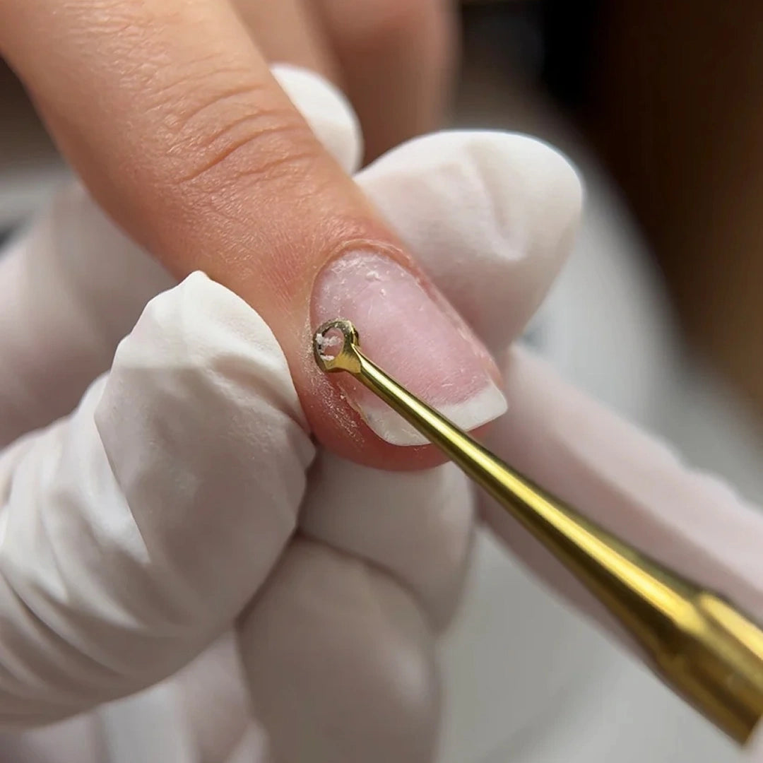 A close-up of a manicurist wearing white gloves using a gold metal cuticle tool to gently lift or clean a fingernail.