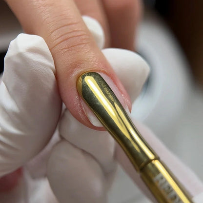 A close-up of a manicure application showing a gold metal double ended cuticle pusher pushing back the cuticle with a white gloved hand holding the finger.